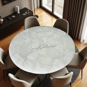 May include: A round, white marble-look dining table with a subtle veined pattern. The table is surrounded by six beige upholstered chairs. The table has the text "Epoxy Agates Boutique" on the surface.