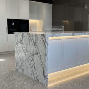 May include: A contemporary kitchen island with a marble worktop and white cupboards. The island has a waterfall edge and is lit with warm LED lighting. The text "EPOXYAGATEBOUTIQUE" is displayed on a clear sign.
