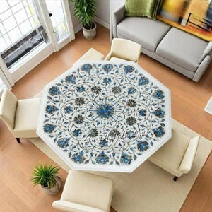 May include: An overhead view of a white octagonal table with a floral design in blue and gray. The table is surrounded by four cream-colored chairs. The room has hardwood floors, a gray sofa, and potted plants.