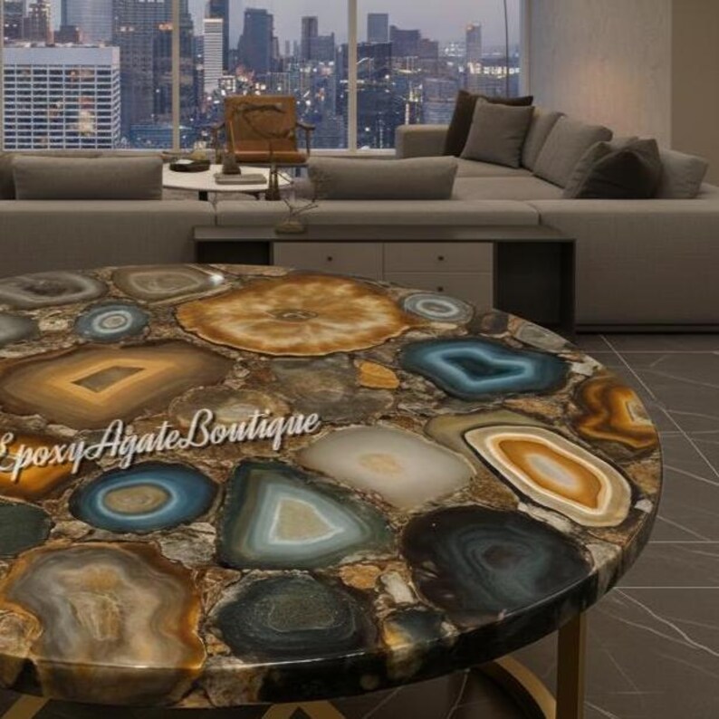 May include: Round coffee table with a unique agate stone top. The table features a mosaic of colorful agate slices in shades of brown, blue, and gold. The base is a gold-colored metal. The text "Epoxy Agate Boutique" is visible on the table.