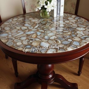 May include: A circular table with a dark wood frame and a polished agate stone top. The tabletop is a mosaic of neutral-toned stones. The table has a pedestal base and is surrounded by two wooden chairs. The table is approximately 76 cm in diameter.
