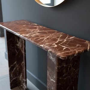 Puede incluir: Una consola rectangular de m&aacute;rmol marr&oacute;n con vetas blancas. La mesa tiene un dise&ntilde;o minimalista con dos patas de soporte. Un espejo con marco dorado cuelga encima, contra una pared gris oscuro.