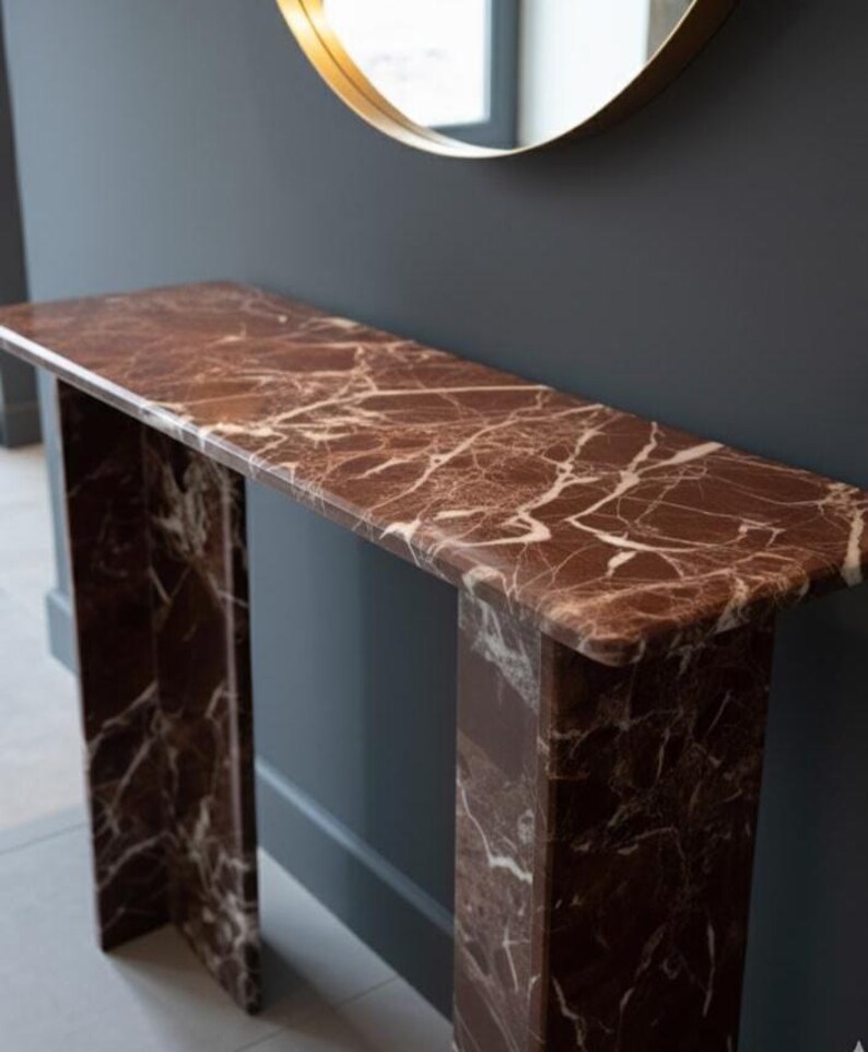 Puede incluir: Una consola rectangular de m&aacute;rmol marr&oacute;n con vetas blancas. La mesa tiene un dise&ntilde;o minimalista con dos patas de soporte. Un espejo con marco dorado cuelga encima, contra una pared gris oscuro.