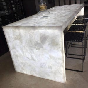May include: A modern, illuminated white quartz countertop with a geometric design. The countertop is paired with black metal stools. A clear glass bottle with a gold accent sits on the counter. The overall design is sleek and contemporary.