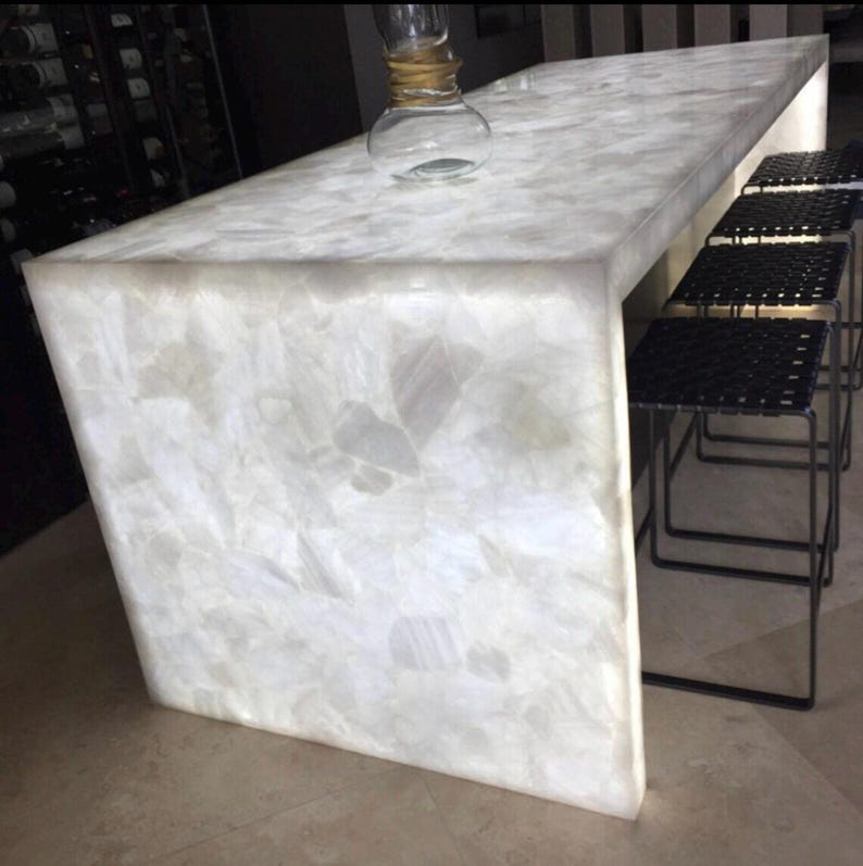 May include: A modern, illuminated white quartz countertop with a geometric design. The countertop is paired with black metal stools. A clear glass bottle with a gold accent sits on the counter. The overall design is sleek and contemporary.