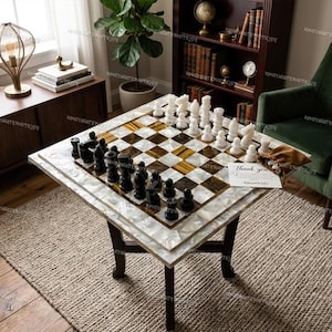 Puede incluir: Una mesa de ajedrez cuadrada con un borde de nácar y una superficie de juego a cuadros. Las piezas de ajedrez son blancas y negras. La mesa está sobre un marco de madera marrón oscuro. Hay una tarjeta de agradecimiento en la mesa.