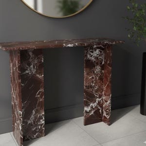 Puede incluir: Una consola rectangular de m&aacute;rmol rojo con vetas blancas. La mesa tiene una parte superior plana y dos patas rectangulares. La mesa est&aacute; colocada contra una pared gris oscuro, con un espejo con marco dorado y un jarr&oacute;n negro con vegetaci&oacute;n en el fondo. Mide unos 120 cm de largo.