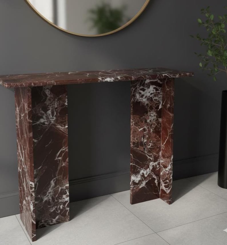 Puede incluir: Una consola rectangular de m&aacute;rmol rojo con vetas blancas. La mesa tiene una parte superior plana y dos patas rectangulares. La mesa est&aacute; colocada contra una pared gris oscuro, con un espejo con marco dorado y un jarr&oacute;n negro con vegetaci&oacute;n en el fondo. Mide unos 120 cm de largo.