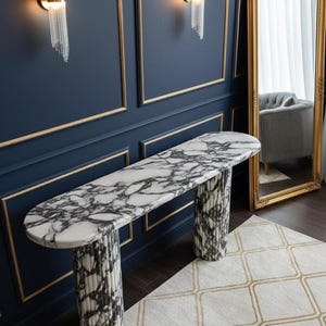 Puede incluir: Una consola con tapa de mármol con un patrón en blanco y negro, sostenida por dos patas cilíndricas estampadas. La mesa está colocada contra una pared azul marino con molduras doradas y apliques de pared. Un espejo con marco dorado refleja un sillón gris.