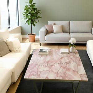 May include: A square coffee table with a pink agate-style surface and a metal frame. The table has the text "EPOXYAGATEBOUTIQUE" on the surface. The table is in a living room with white sofas, a green wall, and a potted plant.