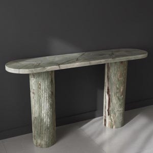 Puede incluir: Una consola de mármol verde con una tapa ovalada y dos patas cilíndricas estriadas. La mesa tiene un aspecto de piedra natural con variaciones de color y veteado. La mesa mide aproximadamente 152 cm de largo.