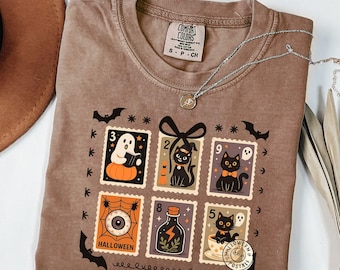 May include: Brown Comfort Colors t-shirt with a Halloween-themed stamp design. The stamps feature a ghost, black cats, an eyeball, and a potion bottle. The shirt has the text "HALLOWEEN POSTALE" and "COMFORT COLORS". A silver necklace is draped across the shirt.