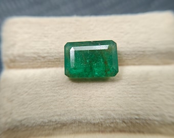 May include: A rectangular emerald gemstone with a vibrant green hue. The gemstone is set against a soft, off-white background, showcasing its clarity and brilliance. The emerald cut highlights the stone's facets.