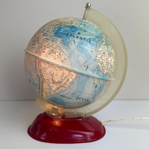 Peut inclure: Une lampe globe vintage avec une base rouge et un cadre en plastique transparent. Le globe représente une carte du monde avec des océans bleus et des masses terrestres beiges. La lampe est éclairée de l'intérieur, illuminant la carte.