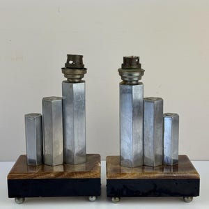 Puede incluir: Par de lámparas de mesa de estilo Art Déco. Cada lámpara presenta un diseño geométrico plateado con tres columnas verticales sobre una base rectangular. La base es una combinación de marrón y negro, con pequeños pies plateados. Los portalámparas son visibles en la parte superior.