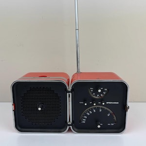 Könnte beinhalten: Ein rotes und schwarzes Retro-Radio mit einer silbernen Antenne. Das Radio hat ein quadratisches Design mit einem Lautsprecher auf der einen Seite und Zifferblättern und einem Abstimmungsanzeiger auf der anderen Seite. Das Radio ist geöffnet und zeigt seine Innenteile.