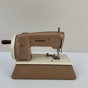 May include: A vintage Singer sewing machine in tan and cream. The machine features a brown hand crank and a cream-coloured base. The Singer logo is visible on the top. The words "Great Britain" are printed on the side.
