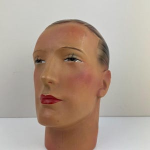 May include: A vintage mannequin head with a painted face. The head has red lips, rosy cheeks, and dark eyelashes. The hair is styled with a side part and is dark brown. The mannequin is made of a light-colored material.