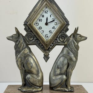 Può includere: Un orologio in stile Art Déco con quadrante quadrato, numeri e lancette neri, incorniciato in una cornice a forma di diamante. Due sculture di cani seduti affiancano l'orologio, il tutto su una base rettangolare in marmo. L'orologio segna l'1:07.