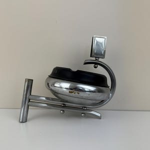 May include: A chrome ashtray with a black rubber insert. The ashtray has a unique design with a rectangular accent and a curved arm. The ashtray is mounted on a metal base and is set against a white background.