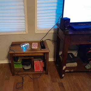 Peut inclure: Une petite table en bois avec une manette de jeu lumineuse et une carte rose. Une console de jeu noire est dans une zone encastrée. La table contient des cartouches de jeu et d'autres appareils électroniques. Une table plus grande avec une télévision et un haut-parleur est en arrière-plan.