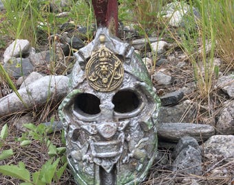 May include: A weathered, silver-colored Roman helmet with a red crest, set amidst rocks and greenery. The helmet features a gold emblem with a lion's head and intricate detailing, showcasing a historical or fantasy theme.