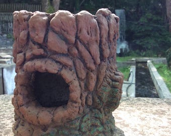 Puede incluir: Una maceta marrón texturizada con forma de tocón de árbol con una gran abertura. La maceta tiene un aspecto rugoso, similar a la piedra, con detalles verdes en la base. El fondo muestra un entorno natural al aire libre.