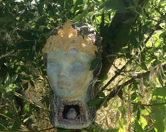 May include: A unique birdhouse shaped like a woman's head, adorned with a golden crown. The face is painted in muted tones, with a small bird nestled inside the opening. The birdhouse is mounted on a tree branch, surrounded by green foliage.