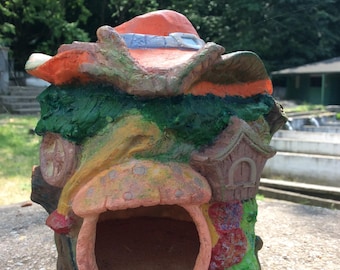 Puede incluir: Una caprichosa pajarera de cerámica pintada a mano con forma de cabaña de fantasía. La pajarera presenta una gran abertura, un tejado con forma de seta y una pequeña casa con una puerta. La paleta de colores incluye naranja, verde y rosa.