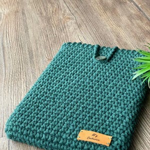 May include: A teal green crocheted tablet sleeve with a button closure. The sleeve has a textured pattern and a small leather tag that reads "A'z Exclusive".