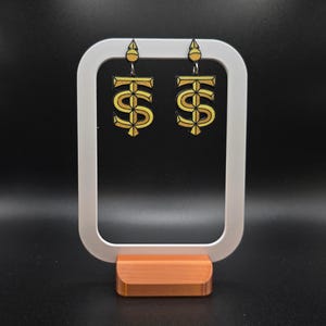 Op de afbeelding: Goudkleurige oorbellen in de vorm van een gestileerd dollarteken, hangend aan een klein goudkleurig topstuk. De oorbellen worden gepresenteerd op een witte en oranje standaard tegen een zwarte achtergrond.