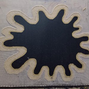Puede incluir: Un lienzo cuadrado de color gris claro con una gran forma negra irregular en el centro. La forma negra est&aacute; delineada con un borde grueso de color crema con un detalle de hilo dorado. El lienzo est&aacute; cosido alrededor de los bordes.