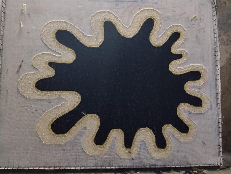 Puede incluir: Un lienzo cuadrado de color gris claro con una gran forma negra irregular en el centro. La forma negra est&aacute; delineada con un borde grueso de color crema con un detalle de hilo dorado. El lienzo est&aacute; cosido alrededor de los bordes.