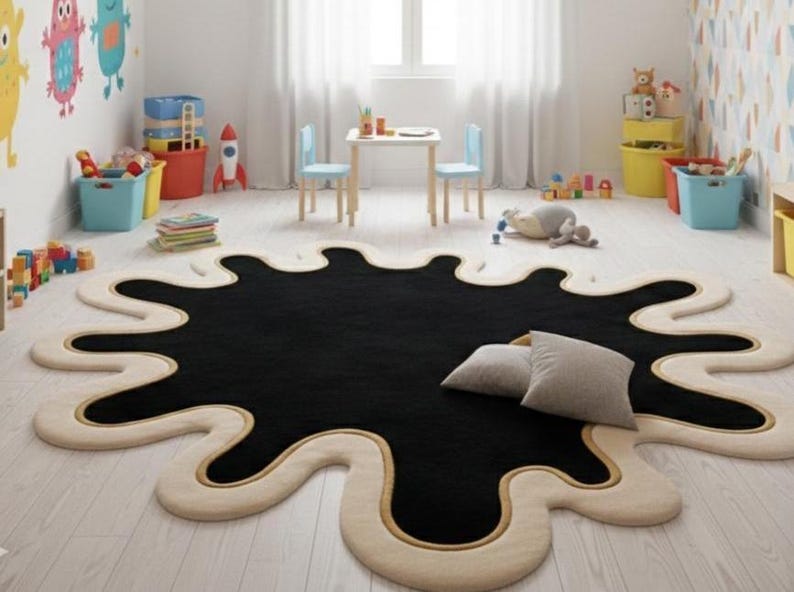 Puede incluir: Una alfombra negra grande con una forma irregular que se asemeja a una mancha de tinta. La alfombra tiene un borde color crema con un fino acento dorado. Dos almohadas grises descansan sobre la alfombra en una habitaci&oacute;n infantil.
