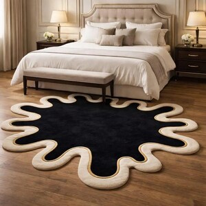 Puede incluir: Una alfombra negra y crema con una forma irregular, similar a una salpicadura. La alfombra tiene un centro negro, un borde crema y una fina l&iacute;nea dorada. La alfombra est&aacute; colocada sobre un suelo de madera en un dormitorio.