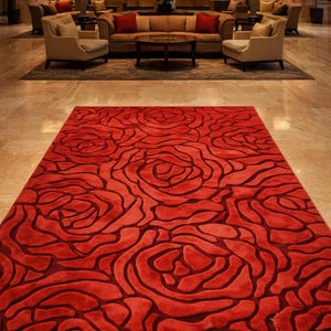 May include: A large, rectangular red rug with a rose pattern in a hotel lobby. The rug's design features dark red outlines that define the floral shapes. The lobby includes seating areas with sofas, chairs, and tables, and is illuminated by warm lighting.