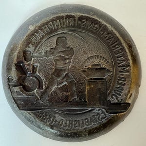 May include: A circular, bronze-coloured metal medallion featuring a relief of a blacksmith. The image includes a forge, anvil, and text that reads "Birmingham Manufacturers" and "Established 1888."