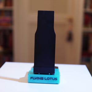 May include: A black, rectangular object with a unique shape, standing on a turquoise base. The base has the text "FLYING LOTUS" in white. The background is blurred, with bookshelves visible.