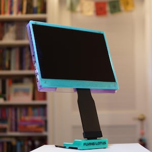 以下が含まれることがあります： 黒い画面のティールと紫色の電子デバイス。黒いスタンドに取り付けられています。スタンドのベースはティールで、「FLYING LOTUS」の文字が印刷されています。デバイスは、本棚のぼやけた背景に配置されています。