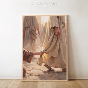 以下が含まれることがあります： GRACE INK ART christian design studioと題された額入りアートプリント。石畳の道を歩く薄いベージュのローブを着た人物と、そのローブの裾に手を伸ばす別の人物が描かれており、背景には他の人影も見えます。