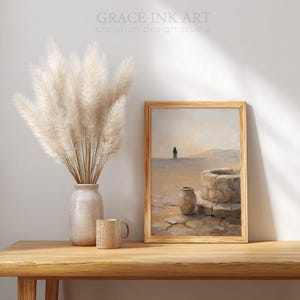 a wooden table with a vase of white flowers, a coffee mug, and a framed painting depicting a desert landscape with a well and a solitary figure.