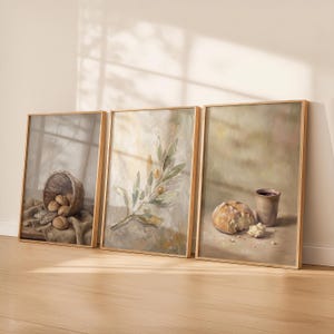 May include: Three framed art prints with a rustic theme. One shows a basket of bread rolls, another an olive branch, and the third a loaf of bread with a cup. The frames are a light brown colour, and the artwork has a muted colour palette.