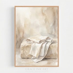 May include: A watercolour print featuring a stone block draped with white cloth, set against a soft, neutral backdrop. The artwork is framed in a light brown wooden frame. The text "GRACE INK ART christian design studio" is at the bottom.