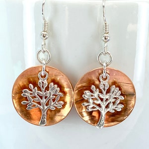 May include: Copper-coloured disc earrings with a silver tree design. The earrings have silver hooks and are suspended from a silver ring. The tree design is a detailed silhouette of a tree with leaves.