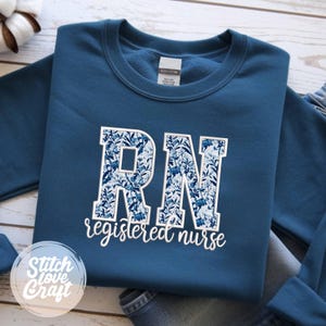 Può includere: Una felpa blu navy con le lettere "RN" in un motivo floreale blu, sopra le parole "registered nurse" in corsivo bianco. La felpa è piegata, con un logo Stitch Love Craft visibile.