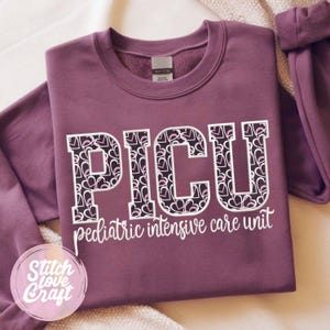 Puede incluir: Una sudadera malva de manga larga y cuello redondo con el texto PICU en letras grandes de bloque rellenas con un patrón de corazones blancos y rosas. Debajo de las letras, el texto pediatric intensive care unit está impreso en letra cursiva blanca.