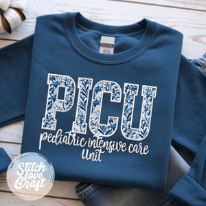 Può includere: Una felpa blu navy con le lettere "PICU" in un motivo floreale bianco. Sotto, le parole "pediatric intensive care unit" sono stampate in bianco. La felpa ha un girocollo e maniche lunghe.