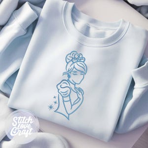 Op de afbeelding: Een lichtblauwe sweatshirt met een blauwe lijntekening van een prinses met een kroon en oorbellen. Het ontwerp bevat een jurk met pofmouwen en de tekst "Stitch Love Craft" in een cirkelvormig logo.