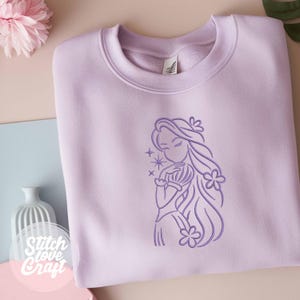 May include: A light purple sweatshirt with a line drawing of a woman with long hair and flowers. The design is in a darker shade of purple. The sweatshirt has a crew neck and long sleeves. The text "Stitch Love Craft" is at the bottom left.