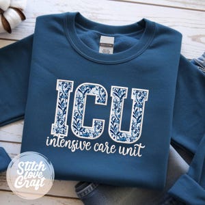 May include: A navy blue sweatshirt with the letters "ICU" in a floral pattern. Below the letters, the words "intensive care unit" are printed in white. The sweatshirt is folded, with a portion of denim jeans visible.